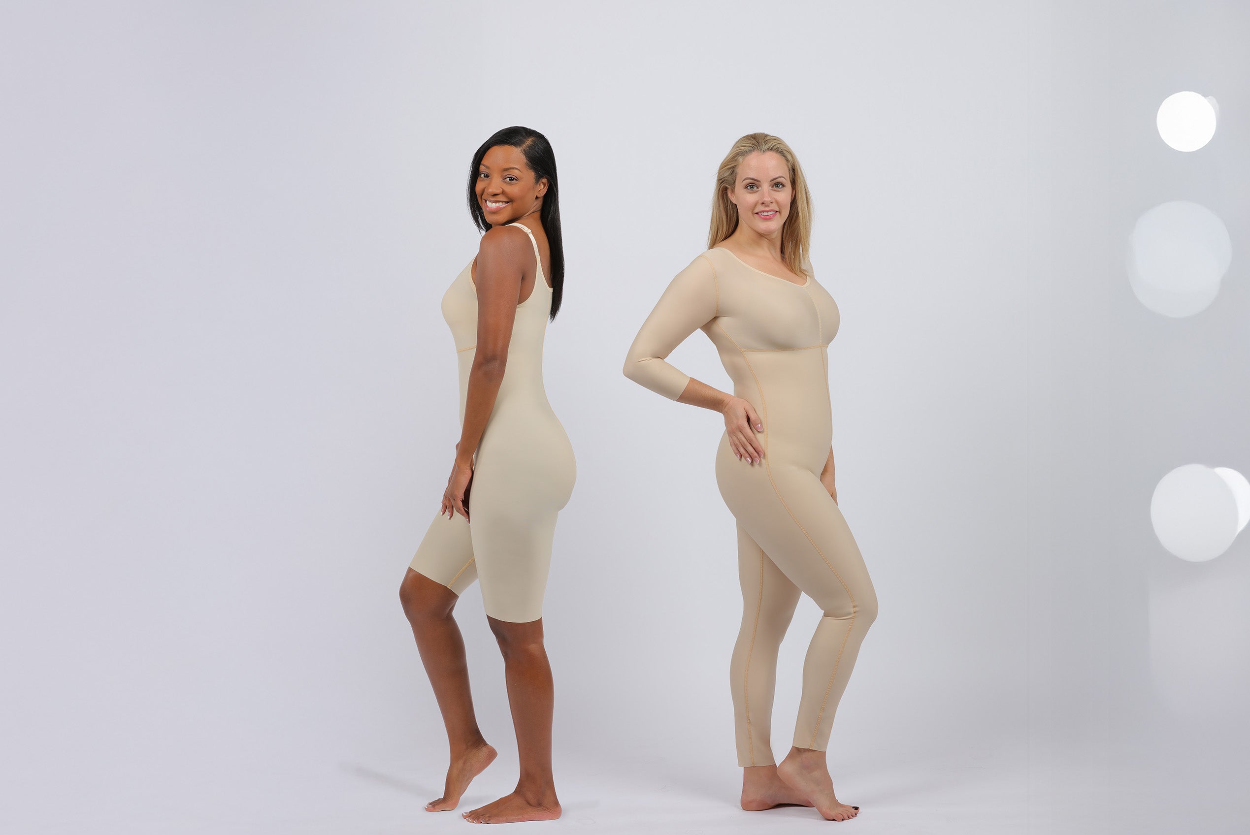 two women posing side by side wearing beige compression bodysuits while facing the camera